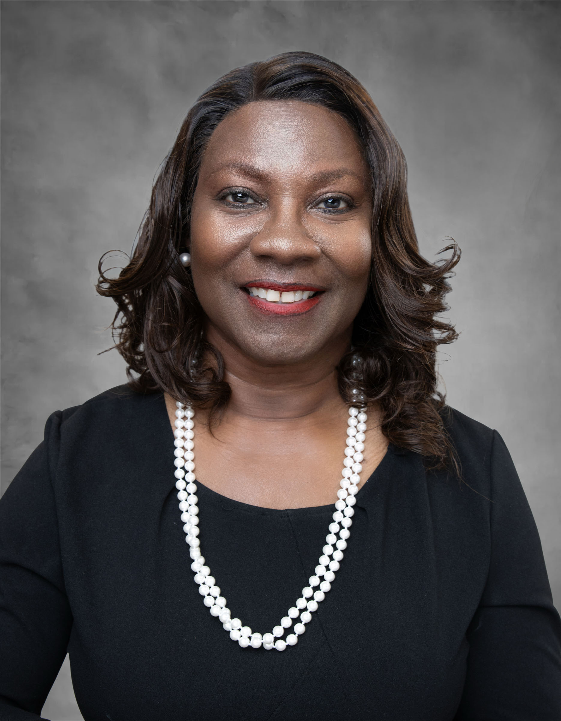 Picture of Executive Director Dr. Delores Dacosta dressed in a black dress with white pearl necklace on grey background.